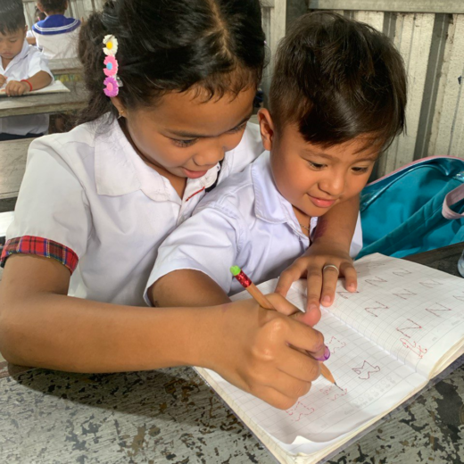 A girl in Cambodia helps a younger classmate with his writing. She has a brightly coloured daisy clip in her hair.