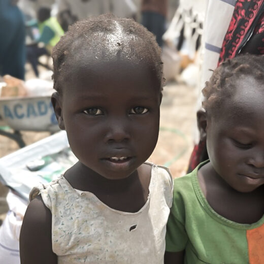 A young girl in South Sudan stares directly at the camera, her white top full of holes. A younger sibling stands next to her in a green and orange top. In the background, we can see the rough ground and a few bags of grain.
