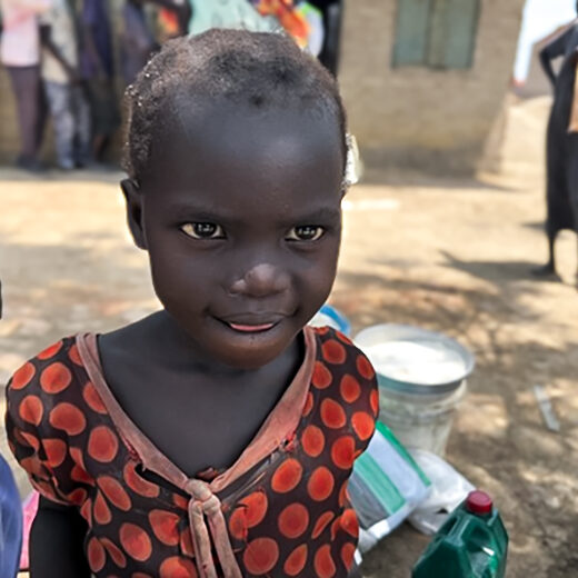 A young girl from South Sudan stares into the camera, half a smile on her face. Her clothes are ragged and dirty.