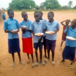 A line of six children in blue school uniform shirts, holding half-eaten plates of food, against a sandy dusty background