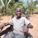 Boy with huge smile seated in a wheelchair against a dusty sandy background with some trees and bushes