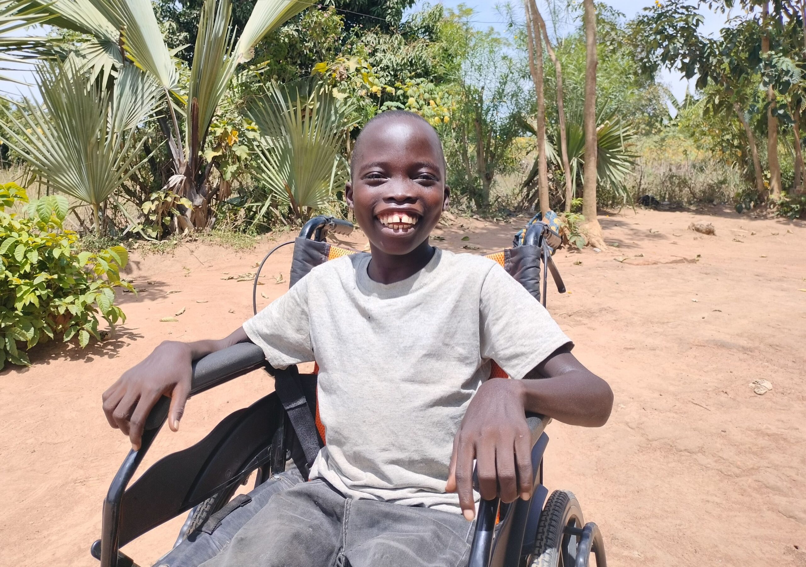 Boy with huge smile seated in a wheelchair against a dusty sandy background with some trees and bushes